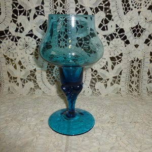 May include: A teal-colored glass candle holder with a rounded top and a flared base. The stem has a decorative design. The candle holder is made of glass and has a glossy finish. The candle holder is a decorative item.
