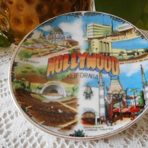 May include: A vintage souvenir plate featuring a colorful illustration of Hollywood, California. The plate depicts iconic landmarks like Grauman's Chinese Theatre, the Hollywood sign, and the Hollywood Bowl. The text "HOLLYWOOD CALIFORNIA" is prominently displayed in a bold, orange font.