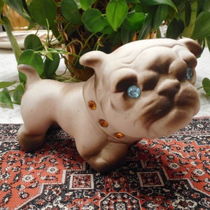 May include: A ceramic figurine of a brown and white bulldog with blue eyes and a jeweled collar. The figurine is sitting on a red and black patterned rug.