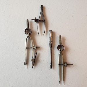 May include: An assortment of vintage drafting tools, including two compasses, a divider, and a screwdriver. The tools are made of metal and appear to be well-used, with a patina suggesting age. The tools are arranged on a light-colored surface.