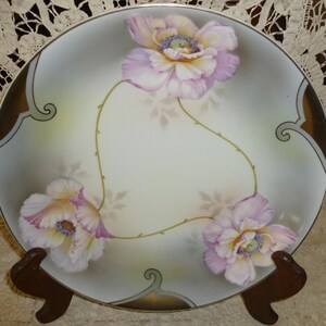 May include: A decorative porcelain plate featuring hand-painted pink and white flowers with yellow centers. The flowers are connected by thin green stems, set against a cream-colored background with gold accents. The plate is displayed on a wooden stand.