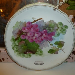 May include: White ceramic plate with a painted design of a bunch of grapes with green leaves. The plate has a gold rim and the text "Compliments of Branson & Hedberg, Cokato, Minn." printed on the bottom.