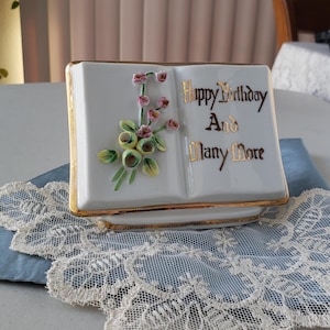 May include: A white porcelain book-shaped trinket box with gold trim and a floral design. The box has the words "Happy Birthday And Many More" written in gold lettering.