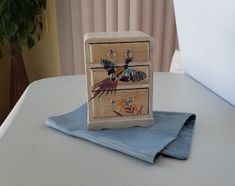May include: A small wooden chest with three drawers, painted white with a decorative design of a pheasant in flight. The drawers have silver knobs.