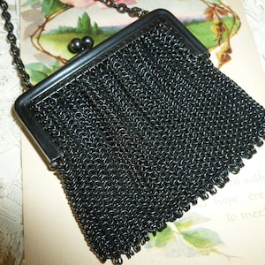 May include: A black metal mesh handbag featuring a rectangular frame with a dual ball clasp closure. The purse is constructed from small interlocking dark metal rings and includes a thin matching black metal chain shoulder strap.