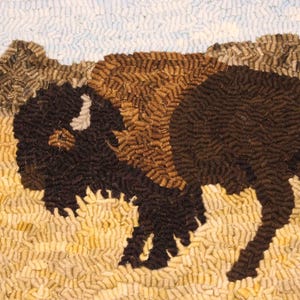 May include: A handcrafted rug featuring a detailed image of a bison. The bison is depicted in shades of brown and black, set against a backdrop of a light yellow field, brown hills, and a light blue sky. The rug's texture is visible.