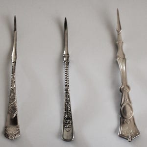 May include: Three antique silver tools with pointed tips. The tools have ornate designs on their handles, including floral patterns and textured details. The tools are displayed against a plain white background, highlighting their intricate craftsmanship.