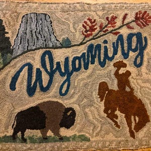 May include: A beige rug with a blue embroidered word "Wyoming" and a brown silhouette of a cowboy riding a horse. The rug also features a brown buffalo, a blue mountain, and a red flower.