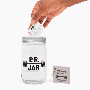 PR Jar™ for Personal Records, Weightlifting, Powerlifting, Crossfit. Jar is clear. Design is black. Coin slot lid is silver. Notepad included. The PR Jar™ is an original FitFizz creation that makes an excellent gift for yourself or any lifter.
