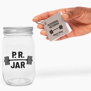 PR Jar™ for Personal Records, Weightlifting, Powerlifting, Crossfit. Jar is clear. Design is black. Coin slot lid is silver. Notepad included. The PR Jar™ is an original FitFizz creation that makes an excellent gift for yourself or any lifter.