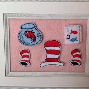 May include: A white frame with a pink background and five embroidered patches. The patches include a fishbowl with a red fish, a red and white striped hat, a book with the numbers 1 and 2 and two fish, and two more red and white striped hats.