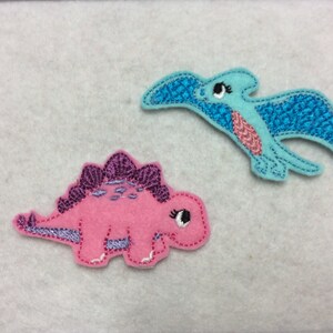 May include: Two felt dinosaur appliques. One is a pink stegosaurus with purple spikes and the other is a light blue pterodactyl with a glittery blue wing.