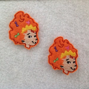 May include: Two felt appliques of a cartoon girl with red hair, a yellow crown, and a colorful bow. The appliques are on a white background.