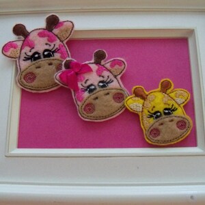 May include: A framed artwork featuring three giraffe head appliqués. Two giraffes are pink with brown spots and accents, one with a pink bow. The third giraffe is yellow with brown spots. The background is a bright pink, and the frame is white.