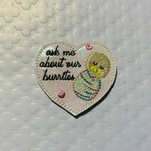 May include: White heart-shaped patch with pink glitter and embroidered text that says "Ask me about our burritos". A small embroidered burrito is in the center of the heart.