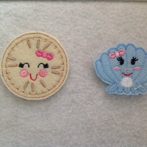 May include: Two felt appliques, one a clock face with a pink bow and the other a blue seashell with a pink bow. Both have embroidered smiling faces.