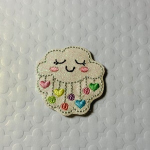 May include: A glittery white cloud-shaped patch with a smiling face, pink cheeks, and closed eyes. Colorful hearts and circles dangle from the cloud. The patch is embroidered with various colors, including pink, yellow, green, and blue.