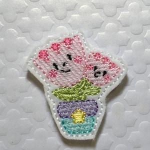 May include: Embroidered patch of two pink flowers with happy faces in a green pot with a purple and blue base and a yellow star.