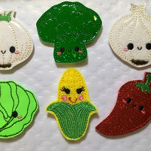 May include: Six felt appliques of different vegetables with cute faces. The vegetables are a green broccoli, a white garlic, a green cabbage, a yellow corn, a green pepper, and a red chili pepper.