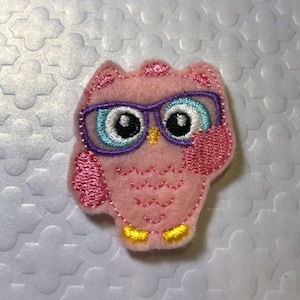 May include: Pink felt owl with blue and purple glasses. The owl has a pink beak and yellow feet.