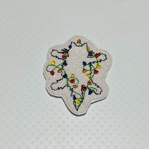 May include: White felt applique of a cat tangled in Christmas lights. The lights are red, green, blue, and yellow.