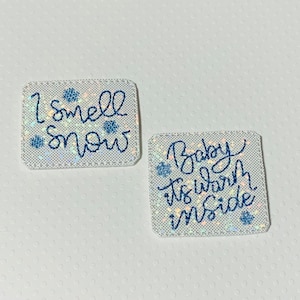 May include: Two white square-shaped fabric patches with blue glitter and blue snowflake designs. The first patch says "I smell snow" and the second patch says "Baby is warm inside".