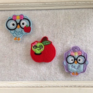 May include: Three felt appliques of owls wearing glasses and holding apples. The owls are blue, purple, and green. The apples are red.