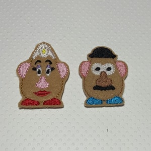 May include: Two felt toy characters, Mr. and Mrs. Potato Head, with embroidered details. Mr. Potato Head has a black hat, brown eyebrows, a brown mustache, and a brown nose. Mrs. Potato Head has a white crown with a yellow star, pink cheeks, and a red mouth.