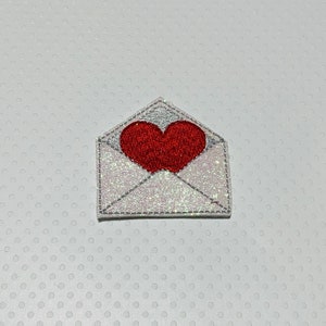 May include: A white envelope with a red heart in the center. The envelope is covered in glitter.