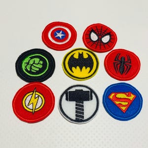 May include: Nine embroidered iron-on patches featuring various superhero logos. The patches are circular and feature the logos for Superman, Batman, Spiderman, The Flash, Thor, Hulk, Captain America, and two different versions of the logo for the character, Spiderman.