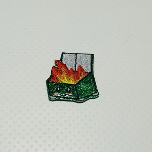 May include: A green glitter-covered trash can with a smiling face and flames coming out of the top. The trash can is a cartoon-style patch.