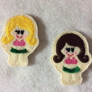 May include: Two embroidered felt figures of girls wearing swimsuits. The girl on the left has blonde hair and is wearing a pink swimsuit with a green skirt. The girl on the right has brown hair and is wearing a pink swimsuit with a green skirt.