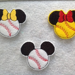 May include: Three felt appliques shaped like Mickey Mouse heads with baseballs for faces. The baseballs are white with red stitching and each applique has a different colored bow on top. One bow is yellow, one is red, and one is black.