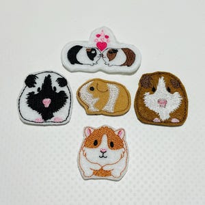 May include: Five felt appliques of guinea pigs. Two are brown and white, one is black and white, one is brown, and one is white with a pink heart above them.
