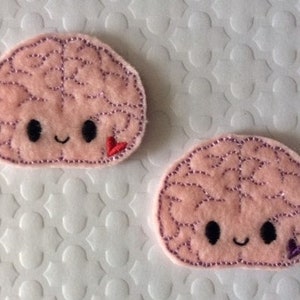 May include: Two pink felt brain-shaped patches with embroidered details. Each brain has a smiling face and a small heart embroidered on the side. The heart on the left brain is red, and the heart on the right brain is purple.
