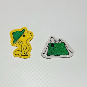May include: Two embroidered patches featuring cartoon characters. One shows a yellow bird with a green hat. The other depicts a white dog and another character inside a green tent with white outlines.