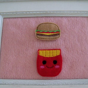 May include: A white frame with a pink background. Inside the frame are two felt appliques: a brown hamburger and a red ketchup bottle with a smiling face.