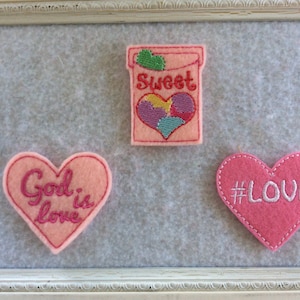 May include: Three felt heart-shaped patches with embroidered designs. The first patch is pink with the text "God is love" in pink. The second patch is pink with the text "#LOVE" in white. The third patch is pink with a box of candy hearts that says "Sweet" and has a heart with multi-colored candy hearts inside.
