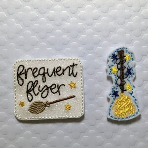 May include: Two white felt patches with gold glitter. One patch says "frequent flyer" with a brown and gold broom. The other patch has a brown and gold broom with blue and gold stars.