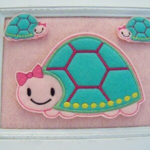 May include: A framed wall decor featuring three felt turtles. The largest turtle is pink with a teal shell and yellow accents. The two smaller turtles are teal with pink accents.