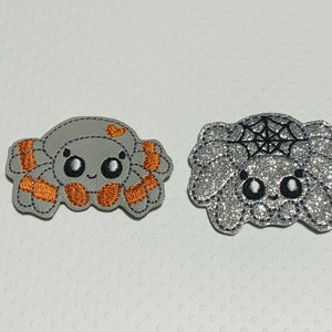 May include: Two embroidered spider patches. One is gray with orange accents, the other is silver glitter with a black spiderweb design. Both have smiling faces and are likely for Halloween.