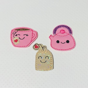 May include: Three pink felt appliques with embroidered faces. A teacup with a heart, a teapot with a purple lid, and a milk carton with a heart.