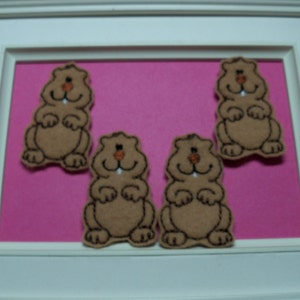 May include: Four brown felt animal appliques shaped like groundhogs with embroidered details. The appliques are on a pink background.