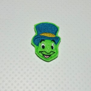 May include: Green felt embroidered patch of Jiminy Cricket from Disney's Pinocchio.  Jiminy is wearing a blue top hat with a gold band.  He has a big smile and is looking to the right.