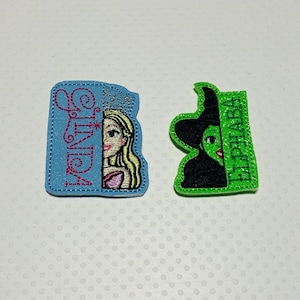 May include: Two embroidered iron-on patches. One is blue with the word "CINDY" and a cartoon image of a girl with blonde hair. The other is green with the word "ELPHABA" and a cartoon image of a woman with green skin and a witch's hat.