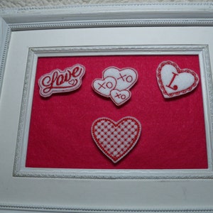 May include: A white picture frame with a red felt background. The frame contains four embroidered hearts with red stitching. One heart says "Love" and another says "XO".