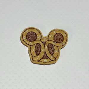 May include: A tan felt applique in the shape of a Mickey Mouse head, with two pretzel shapes forming the ears. The pretzels are brown with white stitching and a brown center.