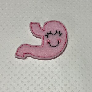 May include: A pink felt stomach with a smiling face and closed eyes, outlined in red stitching. The stomach is a cartoon-like design, with a simple, friendly expression.