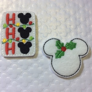 May include: Two felt Christmas ornaments featuring a Mickey Mouse design. One ornament is red, white, and black with the text "Ho Ho Ho" and a string of lights. The other ornament is white with a green holly sprig.