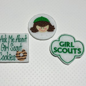 May include: Three embroidered patches for Girl Scouts. One patch features a girl scout in a green hat, another patch says "Girl Scouts", and the third patch says "Ask Me About Girl Scout Cookies" with a chocolate cookie graphic.